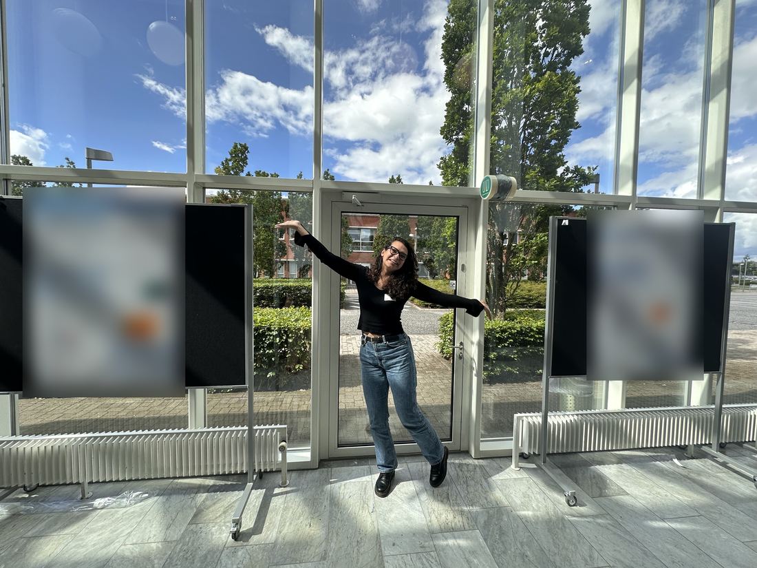 A lab member strikes a joyful pose between two posters in a sunny glass hall at the poster session.